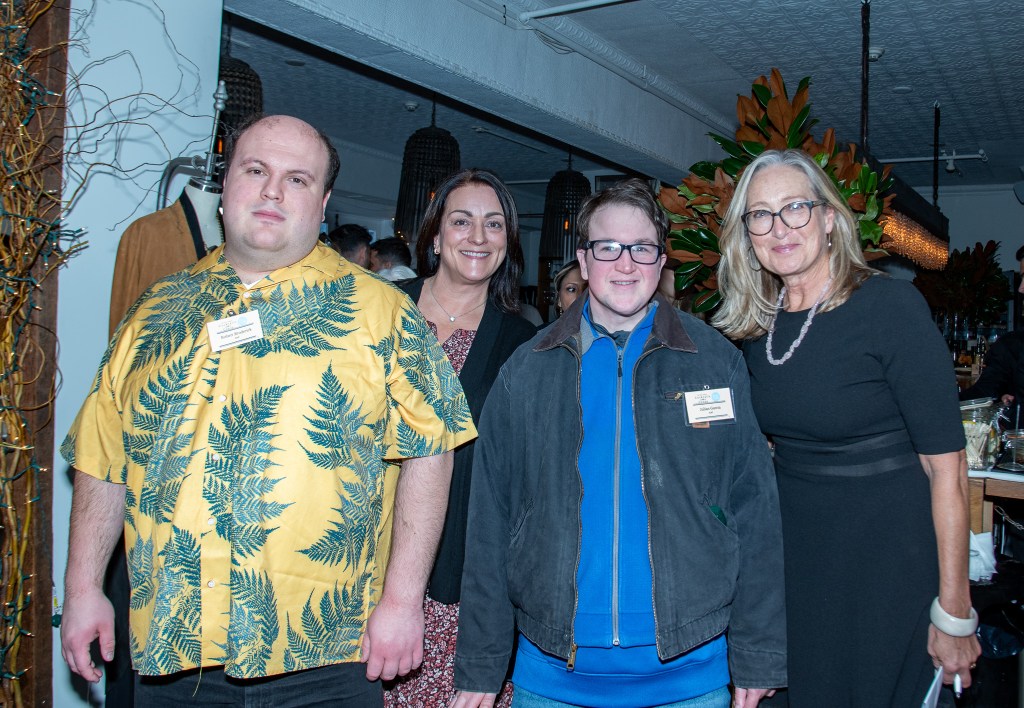 A group of four individuals poses for a photo at the South Fork Bakery's 4th Annual Cocktails for a Cause fundraiser. The scene is lively, showcasing a mix of casual and formal attire, with a backdrop of festive decorations.