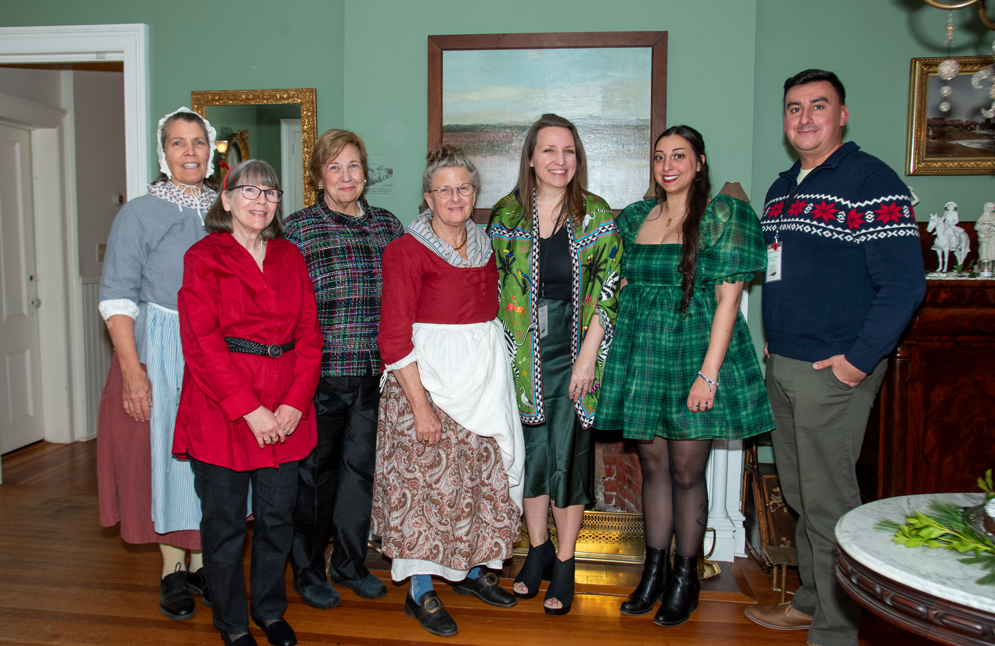 Group of seven people standing together in festive attire, with a tasteful interior background featuring green walls and artwork.