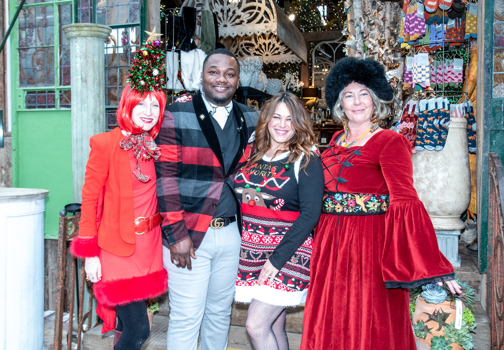 Four people in festive holiday attire posing together in front of a decorative shop entrance, adorned with lights and various items for sale.