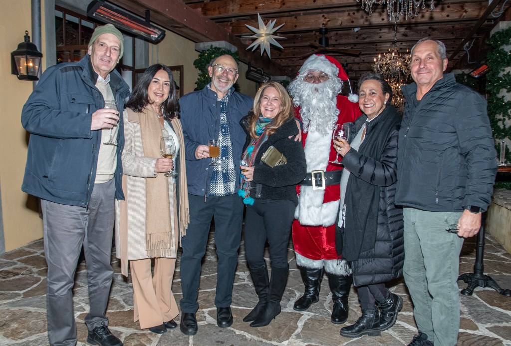 A group of six people gathered together at a festive event, smiling and holding drinks, with a person dressed as Santa Claus among them. The background features a rustic setting with decorative lighting.