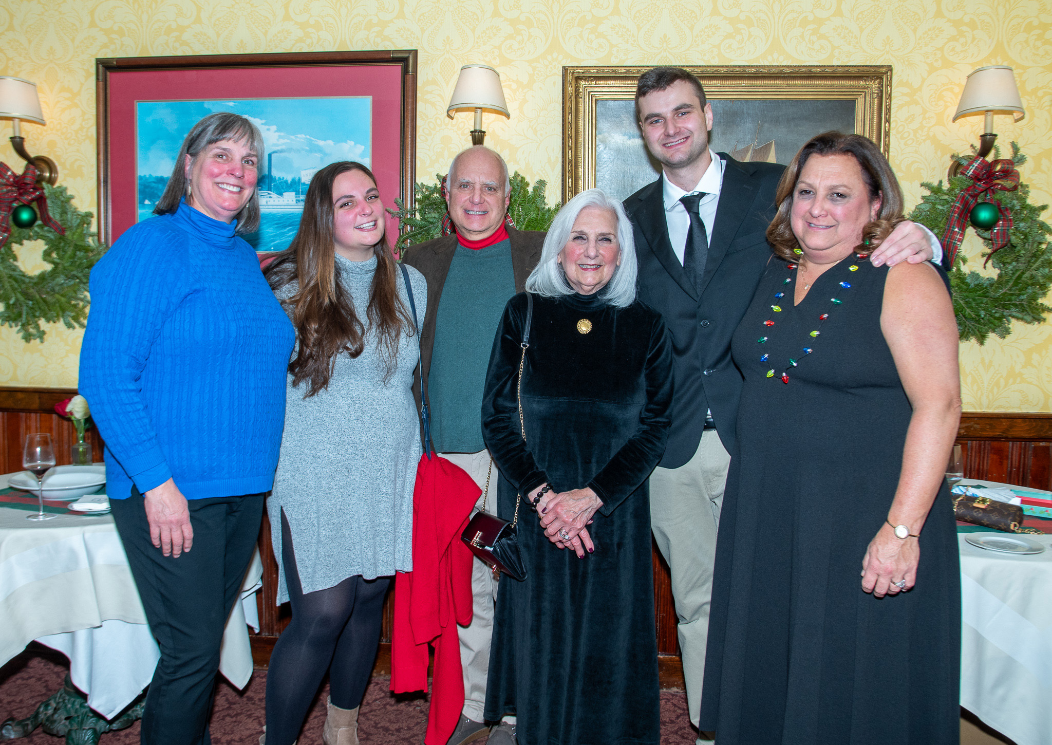 A group of six people smiling together at a festive gathering, adorned with holiday decorations in the background.